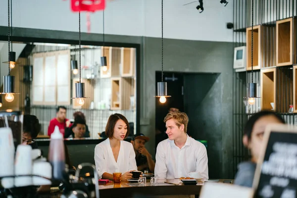 Chinese Asian woman is having coffee with a young Caucasian man. They are catching up casually in the day in a cafe or coworking space and are smiling as they talk comfortably.