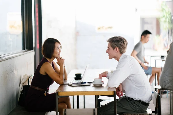 An Asian Chinese woman interviews a young, Caucasian man. She is looking a her laptop computer as she talks with him and they are both smiling. They are sitting at a table in the day in an office.