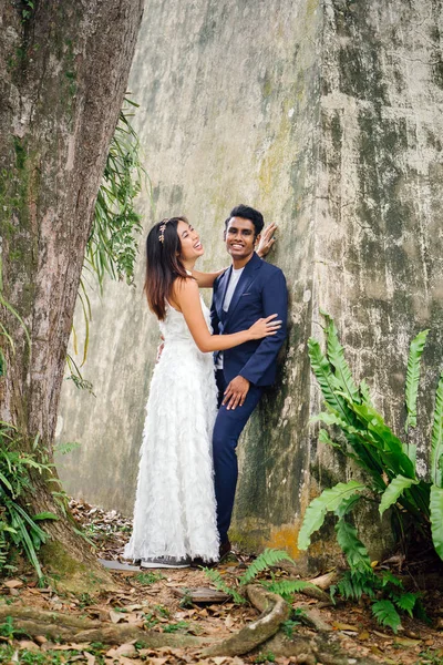 engaged interracial couple (Indian man and Chinese woman) pose for wedding photographs in a park. She is wearing a white wedding dress and he in a blue suit. They are hugging and smiling in joy.