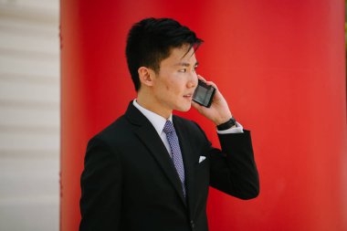 Bir telefon görüşmesi telefonundan ve görüntüleyiciyi gülümseyen Çinli Asyalı adamın portresi. Profesyonel bir kravat ve elbise saat karşı düz kırmızı bir arka plan ile bir takım elbise içinde giyinmiş.