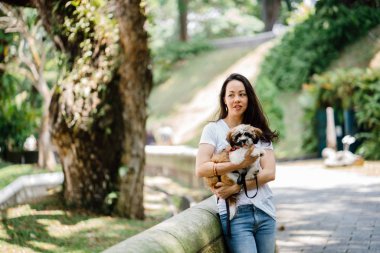 Pet shih tzuh köpeğini gün yeşilliklerle dolu bir parkta ve bitkiler içinde belgili tanımlık geçmiş olan bir genç, çekici ve güzel pan Asya kadın portresi.