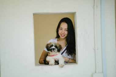 Genç ve çekici genç bir Pan Asya kızla evde beslenen hayvan shih tzuh köpeğini portresi. O gülümseyen ve köpeğini holding ve gün içinde kare bir pencereden bakmak.