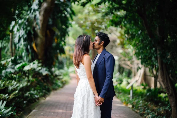 Portrait of interracial couple taking wedding photos in a beautiful park in the day. An Indian man and his Chinese fiance are passionately kissing as they hold hands on a pathway.
