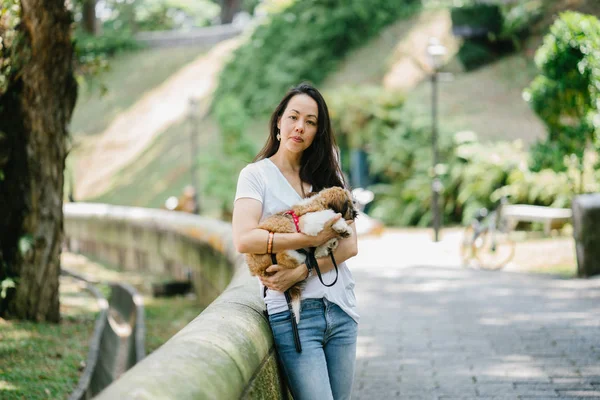 Portrait of a young, attractive pan Asian woman with her shih tzuh dog in the park. They are in the middle of a walk and they look cheerful and relaxed.