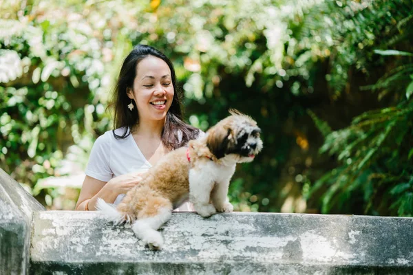 Portrait of a young, attractive pan Asian woman with her shih tzuh dog in the park. They are in the middle of a walk and they look cheerful and relaxed.