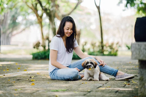 Portrait of a young Pan Asian woman with dog in a green park on a warm day in the park. The dog is a toy breed shih tzuh.