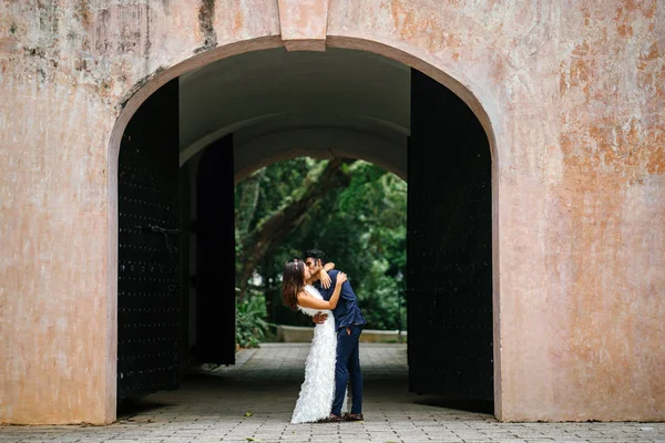 newly engaged couple hug and kiss beneath an arch in a park in Asia. An Indian man and his Chinese wife are taking wedding photographs and are passionately in love with one another.