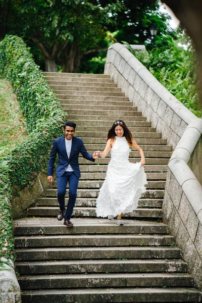 一对跨种族的恋人在公园里跑下来的石头台阶在白天 一个印度男人正和一个中国女人结婚 他们高兴地跑下楼梯 — 图库照片