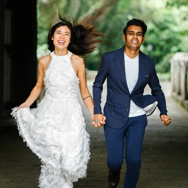 engaged interracial Asian couple run towards the camera in a park in Singapore. An Indian man and his Chinese wife are holding hands and laughing in joy as they run.