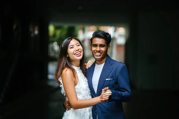 portrait of a young interracial couple getting married. An Indian man and his Chinese wife pose for a photograph in the day along the street against a dark background.