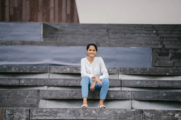 Candid portrait of a smiling attractive, young woman on grey steps . The Indian Asian lady is professional, relaxed and sitting in the bright sun.
