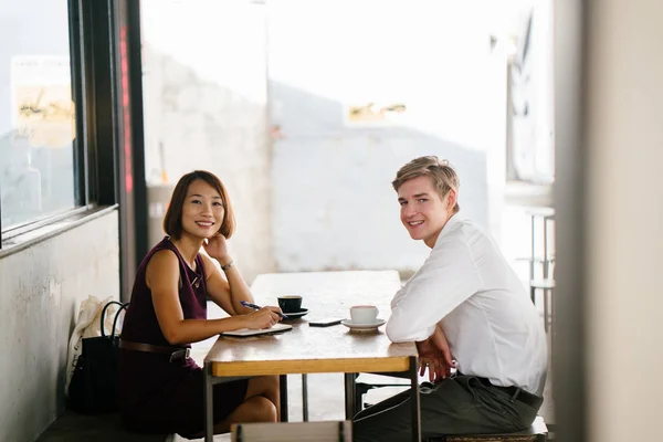 An Asian woman is interviewing a young Caucasian white man for an internship job in Asia. They are sitting at a desk in the shade outside in the day and talking over coffee.