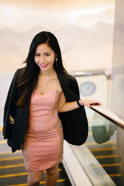 stylish and fashionable young Chinese Asian woman on an escalator. She is wearing a beautiful pink dress with a man\'s jacket draped over her shoulders and smiling.