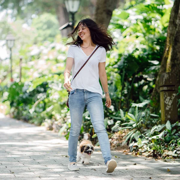 Portrait of a young Pan Asian woman with dog in a green park on a warm day in the park. The dog is a toy breed shih tzuh.