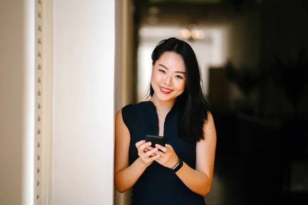 一个年轻迷人的亚洲妇女的肖像 她使用智能手机在一天窗口附近 她穿着一件专业的深蓝色连衣裙 面带微笑 — 图库照片