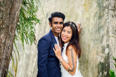 nişanlı ırklararası çift (Hintli ve Çinli kadın) bir parkta düğün fotoğrafları için poz. O giyiyor beyaz gelinlik ve o mavi takım elbiseli. Sarılma ve sevinç içinde gülümseyen.