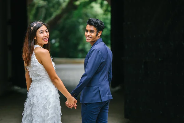 インドの新郎に彼の中国の妻の妻が保持していると公園でサイド サイドのスタンド 異人種間のカップル日のアーチの下の結婚式の写真を取っているし 笑っています — ストック写真