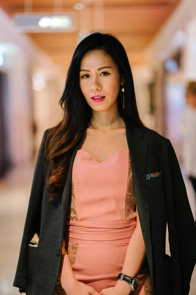 stylish and fashionable young Chinese Asian woman on an escalator. She is wearing a beautiful pink dress with a man's jacket draped over her shoulders and smiling.