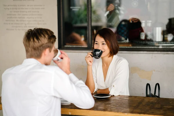 An Asian woman is interviewing a young Caucasian white man for an internship job in Asia. They are sitting at a desk in the shade outside in the day and talking over coffee.