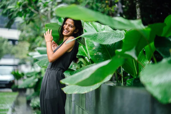 Candid portrait of a young Indian woman standing in a lush green garden. She\'s dressed in a professional grey smock jumpsuit and smiling.
