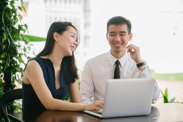 Portrait of two smiling business people (lawyers, consultants, etc) or couple smiling mid discussion over a laptop in an office with greenery in the background. They are focused on the laptop.