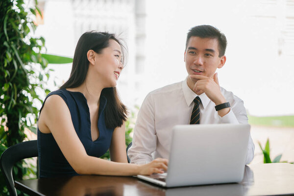 Portrait of two smiling business people (lawyers, consultants, etc) or couple smiling mid discussion over a laptop in an office with greenery in the background. They are focused on the laptop.