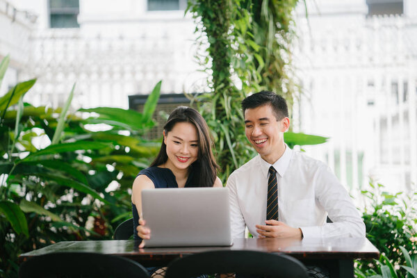 Portrait of two smiling business people (lawyers, consultants, etc) or couple smiling mid discussion over a laptop in an office with greenery in the background. They are focused on the laptop.