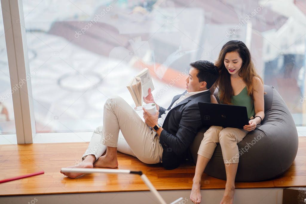 A Chinese Asian couple unwind over the weekend in their apartment. An attractive woman is working on her laptop on a beanbag and her boyfriend is reading a book and leaning against her.