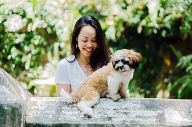 Pet shih tzuh köpeğini gün yeşilliklerle dolu bir parkta ve bitkiler içinde belgili tanımlık geçmiş olan bir genç, çekici ve güzel pan Asya kadın portresi.