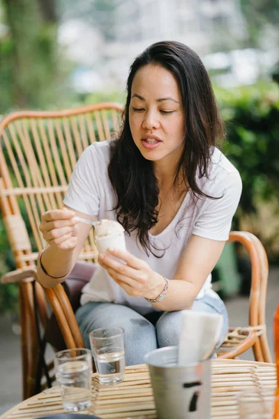 Portrait of a Pan Asian woman relaxing in the sun at a cafe with coffee and ice cream. She is sitting in a rattan chair and smiling while she enjoys the afternoon.