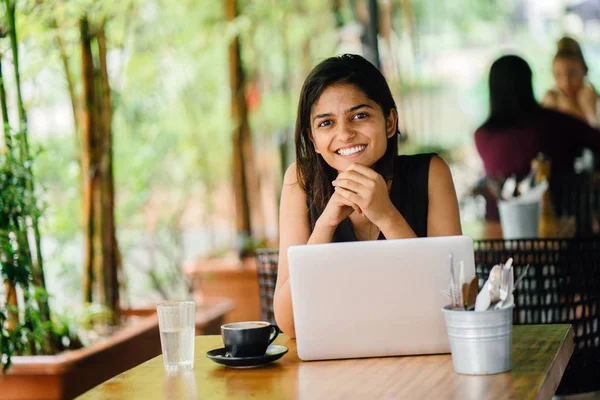 Candid portrait of an attractive and young Indian Asian professional woman work on her laptop in a trendy cafe or coworking space with teal wooden furniture. She is smiling and looking away.