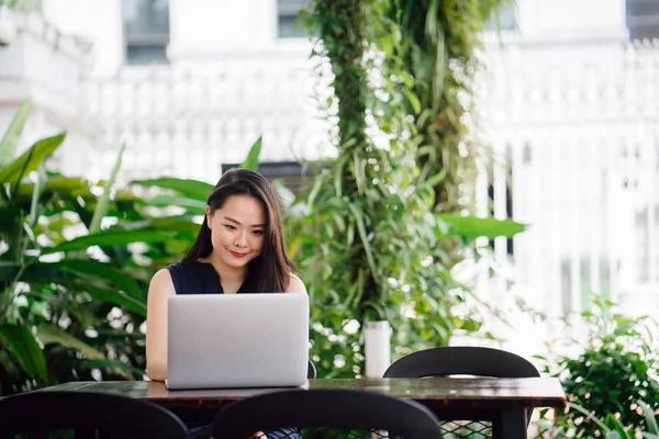 Portrait of a young Chinese Asian woman professional smiling over her laptop in the day. She is seated at the table in an office with green plants behind her.