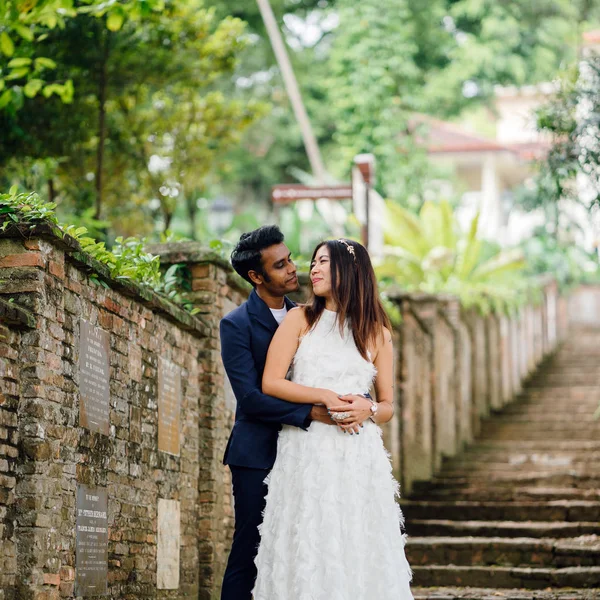 interracial couple (Indian man, Chinese woman) pose for wedding photo in a park in the day. They are on old stone steps and and smiling and laughing as they take photos.