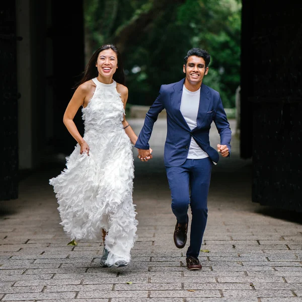 engaged interracial Asian couple run towards the camera in a park in Singapore. An Indian man and his Chinese wife are holding hands and laughing in joy as they run.