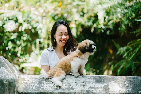 Portrait of a young, attractive pan Asian woman with her shih tzuh dog in the park. They are in the middle of a walk and they look cheerful and relaxed.