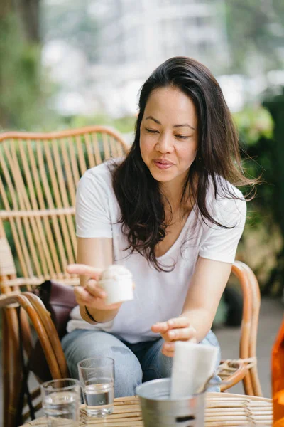 Portrait of a Pan Asian woman relaxing in the sun at a cafe with coffee and ice cream. She is sitting in a rattan chair and smiling while she enjoys the afternoon.