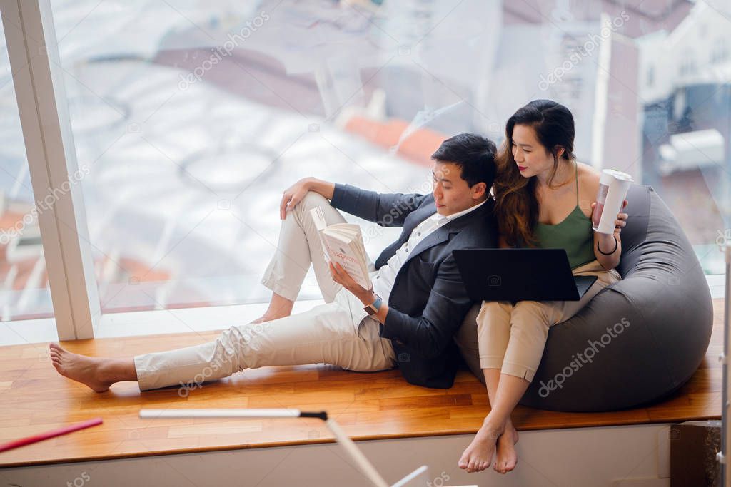 A Chinese Asian couple unwind over the weekend in their apartment. An attractive woman is working on her laptop on a beanbag and her boyfriend is reading a book and leaning against her.