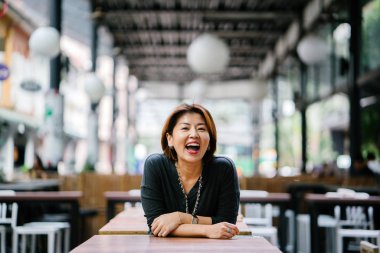 Portrait of an elegant mature Asian woman laughing during the day. She's dressed professionally and leaning against a table in a coworking space, cafe or office in Asia.