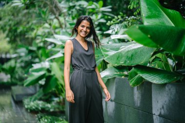 Yemyeşil bir yeşil bahçesinde duran genç Hintli kadın samimi portresi. O bir profesyonel gri giyinmiş senide tulum ve gülümseyen.