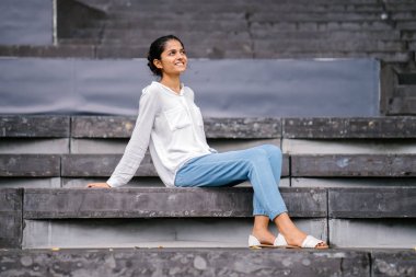 Gri adımlar üzerinde gülümseyen bir çekici, genç kadın samimi portresi. Profesyonel, rahat ve parlak güneşin oturan Hint Asya Bayan.