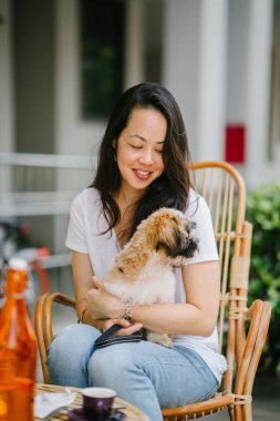  Genç Pan Asya kadın onu hafta sonu evde beslenen hayvan shih tzuh köpeğiyle sahiptir. Köpeğiyle rattan sandalye güneşin bir kafede oturuyor 