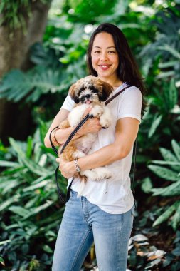 Pet shih tzuh köpeğini gün yeşilliklerle dolu bir parkta ve bitkiler içinde belgili tanımlık geçmiş olan bir genç, çekici ve güzel pan Asya kadın portresi.