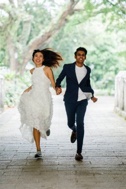  Singapur bir parkta kameraya doğru nişanlı Interracial Asya çift çalıştırın. Bir Hintli ve Çin eşi elele ve sevinç içinde çalışırken gülüyor.