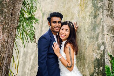 nişanlı ırklararası çift (Hintli ve Çinli kadın) bir parkta düğün fotoğrafları için poz. O giyiyor beyaz gelinlik ve o mavi takım elbiseli. Sarılma ve sevinç içinde gülümseyen.