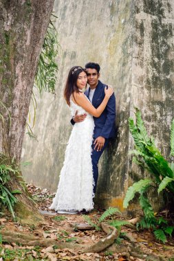 nişanlı ırklararası çift (Hintli ve Çinli kadın) bir parkta düğün fotoğrafları için poz. O giyiyor beyaz gelinlik ve o mavi takım elbiseli. Sarılma ve sevinç içinde gülümseyen.