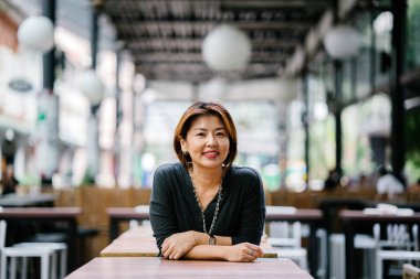 Mature and attractive Asian woman (Korean, Chinese, Japanese) woman leaning against a table in a city in Asia and smiling. She is elegant and professionally dressed.