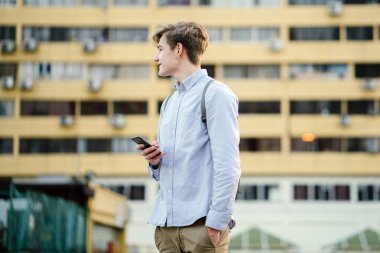 Açık havada duran genç çekici beyaz erkek portresi akıllıca telefon ve gülümseyen ile giyinmiş.