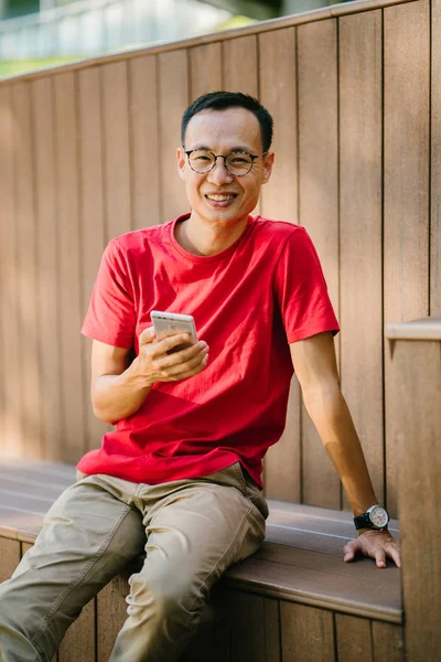 middle-aged Asian man talking on his phone in a park. He is sitting on a wooden structure and smiles as he types on his phone at sunset. He is dressed comfortably in a tee shirt and loose pants.