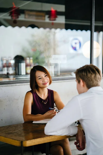 An Asian woman is interviewing a young Caucasian white man for an internship job in Asia. They are sitting at a desk in the shade outside in the day and talking over coffee.