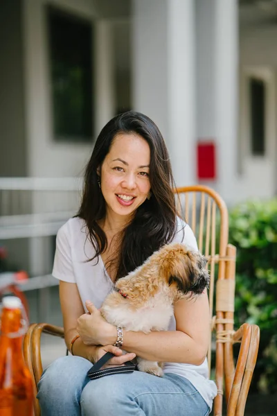 young Pan Asian woman enjoys her weekend with her pet shih tzuh dog. She is sitting in a rattan chair in the sun in a cafe with her dog